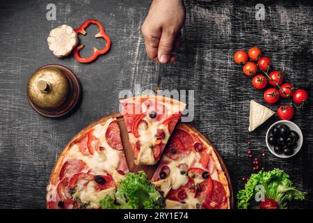 A slice of pizza on a spatula in hand with ham, salami, cheese, mushrooms, cherry tomatoes, bell peppers and greens on a stone and a black chalk board Stock Photo