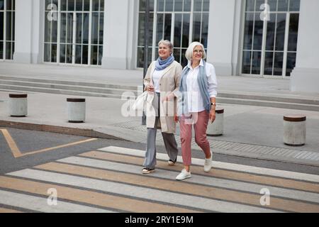 Full length Two elderly women with gray hair elegantly dressed walking, crossing road in city and smiles. Fashionable and stylish senior female friend Stock Photo