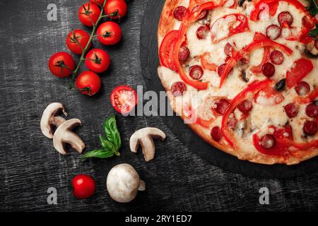 Pizza with smoked sausages, cheese, mushrooms, cherry tomatoes, bell peppers and greens on a stone and a black chalk board, top view. Stock Photo