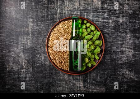 A bottle of beer on a green fresh hop and wheat grain in a plate against the background of a black scratched chalkboard, top view. Stock Photo