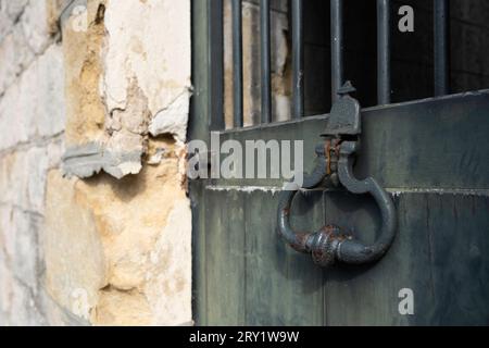 Old closed weathered green metal door with bars on the top and a large antique half-rusted iron door handle in a pale yellow limestone wall Stock Photo