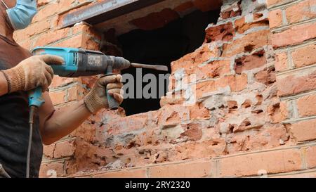 a worker in a black T-shirt and blue jeans with a powerful drill in gloved hands hollows out a hole for a window in a red brick wall from the street Stock Photo