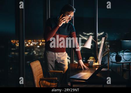 businessman standing at his desk in an office, talking on his mobile phone as he works late into the night. Professional man communicating with his co Stock Photo
