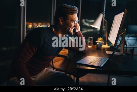 A happy business person works late at night in the office, discussing a project on a phone call. He sits at his desk, focused on his laptop, maintaini Stock Photo