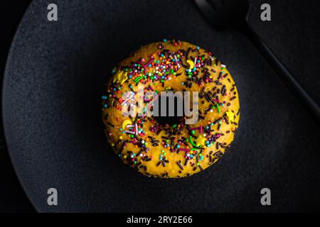 Top view of sweet banana donut with colorful sprinkles placed on black plate against dark surface Stock Photo