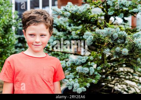 Smiling preteen boy in casual clothes looking away while standing in garden near silver fir tree in daylight Stock Photo