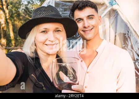 Mature woman in trendy hat having glass of wine while taking selfie with handsome male friend in nature Stock Photo