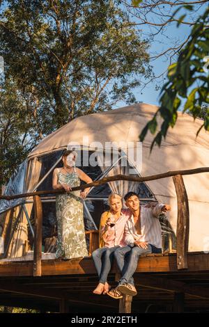 Happy young couple looking away while sitting on wooden structure near tent along with standing mature lady in sunglasses and showing hand away in day Stock Photo