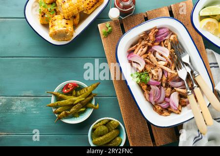 Pulled bbq chicken with baked onions on serving platter, bbq corn, pickles, chili peppers and buns for hot dogs and burgers, tomato sauce. Traditional Stock Photo