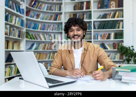 Portrait of a young Indian male student studying in the campus library using a laptop and writing down a lecture in a notebook. He looks at the camera with a smile. Stock Photo