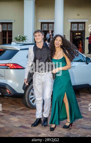 A multiracial couple makes a grand entrance at prom, stepping out in style with a sleek white car behind them, exuding elegance Stock Photo