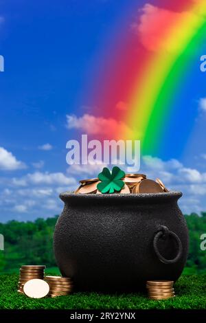 A black kettle pot full of gold coins and a single four leaf clover on top sitting in the grass, there is a rainbow coming out of the top of the pot.  Stock Photo
