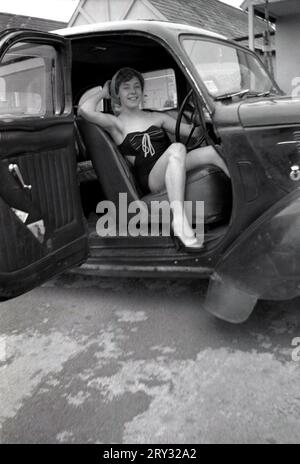 1960s, historical, a young lady in swimsuit sitting in a motorcar, in the drivers seat posing for her photo, outside a chalet at a holiday camp, England, UK. The car has side or rear hinged doors, common on motor cars bullt in the previous era. These were also known as 'suicide' doors, as if the car was moving and they opened, anyone who grabbed the door to stop was thrown out of the vehicle, as seat belts were not in use at that time. Stock Photo