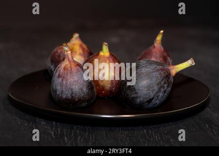 A black plate with five ripe figs placed on a textured black table. Stock Photo