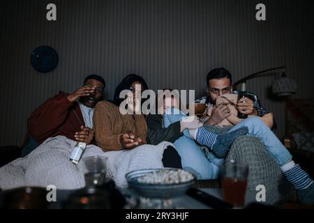 Multiracial friends watching scary movie together while sitting on sofa in cabin Stock Photo