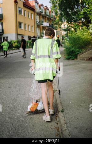 Full length rear view of teenage boy wearing reflective clothing while walking with garbage bag on street Stock Photo