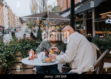 Male and female senior friends discussing over smart phone while having food at sidewalk cafe Stock Photo