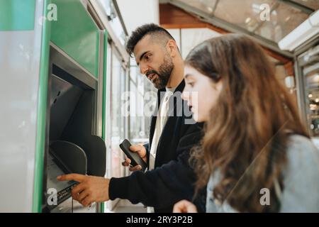 Father using ATM machine while standing with daughter at railroad station Stock Photo