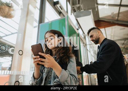 Girl using smart phone with father accessing ATM machine at railroad station Stock Photo