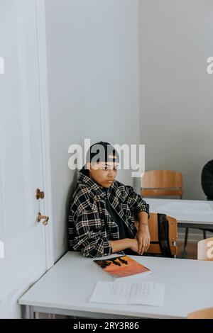 Thoughtful young man sitting at desk while leaning on white wall in classroom Stock Photo