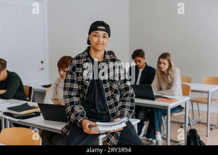 Portrait of smiling young man with book sitting at desk in classroom Stock Photo