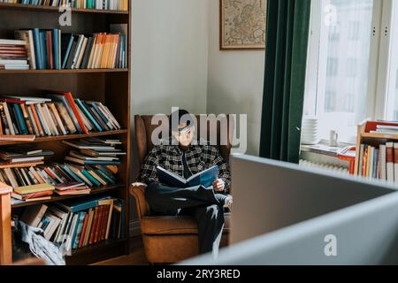 Young man reading book while sitting on armchair by bookshelf in library Stock Photo