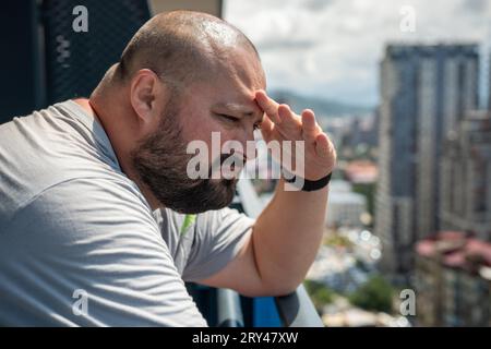 Man covers with hand from sun look into distance from balcony. Fat bald bearded guy hiding with palm Stock Photo