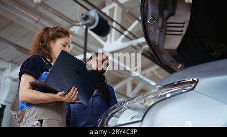 Licensed technician in garage using laptop to follow checklist while doing maintenance on vehicle, talking with client. Worker in car service does checkup on automobile, ground level shot Stock Photo