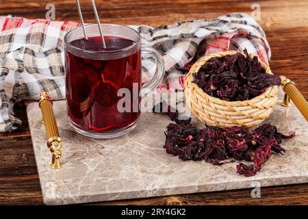 Red hibiscus tea in glass cup on white wooden table with dry rose petals. Hibiscus tea background. Stock Photo