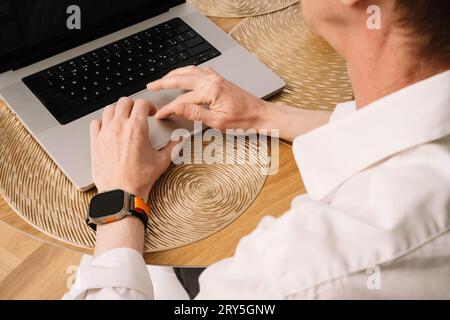 Close up of hands of adult man in white shirt working at laptop at table. Hard-working business man with sporty orange watch on wrist operating on com Stock Photo