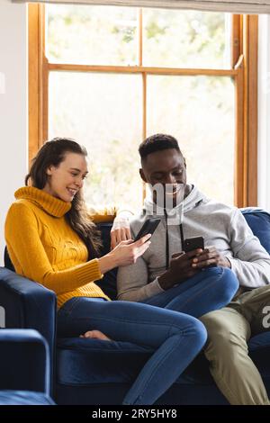 Happy diverse couple sitting on sofa using smartphones at home Stock Photo