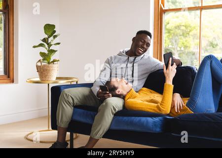 Happy diverse couple sitting on sofa using smartphones at home Stock Photo