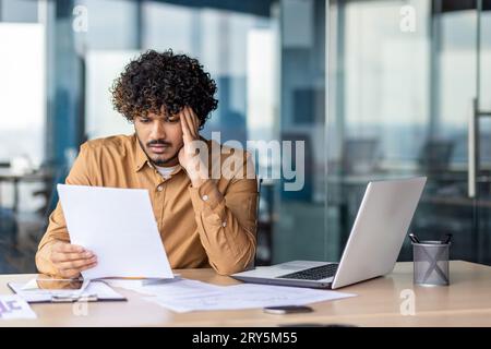 Serious and upset businessman checking and analyzing documents financial reports, thinking hispanic man reviewing contracts on paper work, man inside office at workplace with laptop. Stock Photo