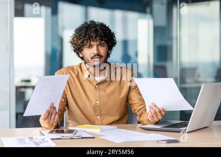 Portrait of young dissatisfied and disappointed financier, businessman behind paperwork looking at camera, negative checking documents and financial reports inside office, using laptop at work. Stock Photo
