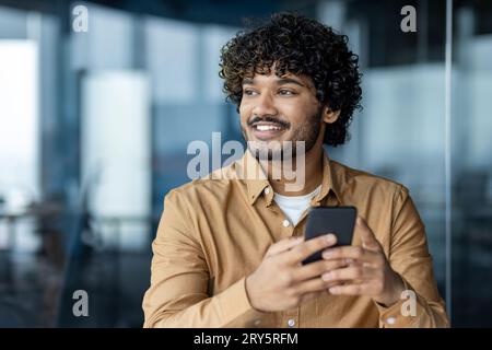 Happy dreamy thinking businessman close-up inside office workplace, hispanic man holding phone, using smartphone app, smiling and looking out window. Stock Photo
