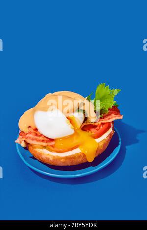 Poached eggs with liquid yolk, tomato, bacon and cheese sauce on toasted bun on plate over blue background. Concept of breakfast, food. Pop art Stock Photo