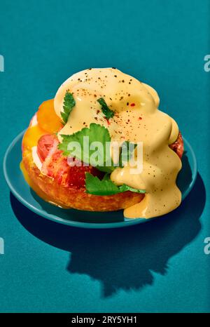 Poached eggs with liquid yolk, tomato, bacon and cheese sauce on toasted bun on plate over cyan background. Concept of breakfast, food. Pop art Stock Photo