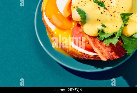 Poached eggs with liquid yolk, tomato, bacon and cheese sauce on toasted bun on plate over cyan background. Concept of breakfast, food. Pop art Stock Photo