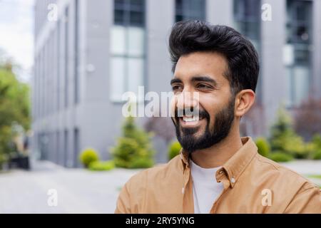 Young dreamy man close-up smiling and looking to the side, portrait of a student businessman in the air outside an office building, in casual clothes. Stock Photo