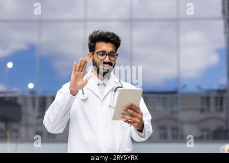 Young male doctor in white medical coat outdoors, smiling and talking consulting patients remotely, doctor using app on tablet computer for video call, waving hand greeting smiling. Stock Photo
