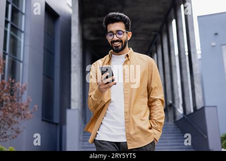 A young smiling man walks through the city with a phone in his hands, outside an office building, happily uses an application on a smartphone, reads messages, types, and browses the Internet. Stock Photo