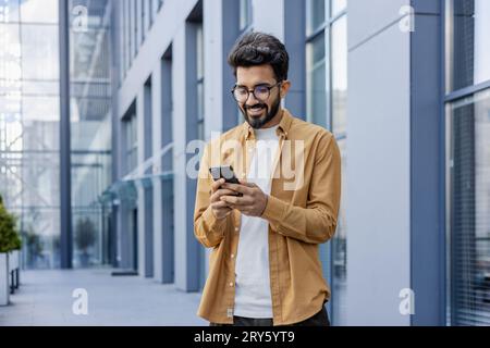 A young smiling man walks through the city with a phone in his hands, outside an office building, happily uses an application on a smartphone, reads messages, types, and browses the Internet. Stock Photo