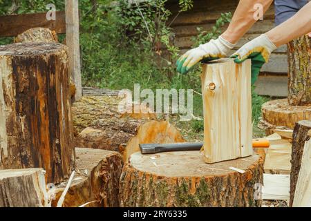 Unrecognizable adult man in gloves standing near countryside house and separating wood on halves with hands after cutting firewood with ax. Tool lying Stock Photo