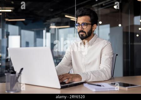 Serious young Indian businessman man working focused on laptop while sitting in office at desk. Stock Photo
