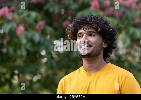 Close-up photo. Portrait of a young Indian man standing in an orange t-shirt outside in a park and looking to the side smiling. Stock Photo