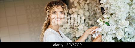 cheerful florist looking at camera while working with white floral decor in event hall, banner Stock Photo