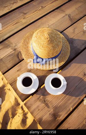 A hat and two cups of coffee stand on a pier near a sandy beach. Stock Photo