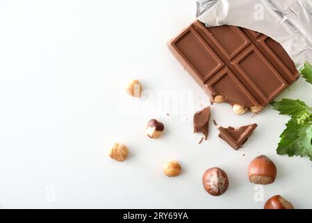 Detail of milk chocolate bar with hazelnuts in aluminum wrapper on white table with nuts and leaves around. Top view. Stock Photo