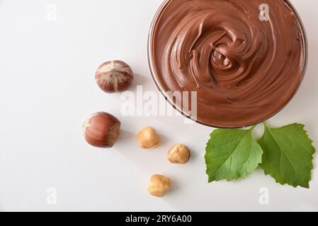 Closeup of chocolate cream with hazelnuts in a bowl isolated on white table with leaves and nuts. Top view. Stock Photo
