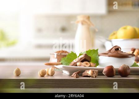 Slices and cream of milk chocolate with hazelnuts on wooden table with leaves and nuts around in a kitchen. Front view. Stock Photo